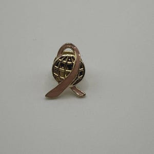 May include: A gold-toned pin featuring a globe design encircled by a pink ribbon. The globe has a grid pattern, and the ribbon is smooth and curved. The pin is set against a plain white background.