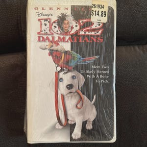 Puede incluir: Cinta VHS sellada de 102 Dálmatas de Disney. La portada presenta a Cruella de Vil, un loro y un cachorro con una correa roja. El título está en rojo y blanco, con un fondo en blanco y negro. El texto dice "Meet Two Unlikely Heroes With A Bone To Pick."