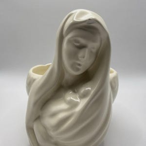 Könnte beinhalten: Cremefarbener Keramik-Blumentopf in Form einer Frau mit drapierter Kopfbedeckung, die eine kleine Figur hält. Der Topf hat einen ruhigen Ausdruck und eine glatte, glänzende Oberfläche. Das Design ist religiös oder spirituell.
