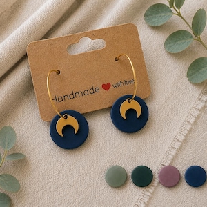 Könnte beinhalten: Ein Paar Creolen mit dunkelblauen runden Anhängern und goldfarbenen Mondsichel-Charms. Die Ohrringe sind auf einem braunen Pappschild mit dem Text Handmade with love und einem roten Herzsymbol befestigt. Fünf weitere runde Anhänger in Salbeigrün, Dunkelgrün, Mauve, Dunkelblau und Schwarz sind darunter abgebildet.