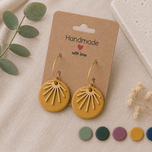 Könnte beinhalten: Ein Paar senfgelbe, kreisförmige Ohrringe mit goldenen Sonnenstrahl-Ausschnitten an dünnen goldenen Creolen, befestigt an einer braunen Pappkarte mit der Aufschrift Handmade with love, ergänzt durch fünf kleine kreisförmige Farbmuster.