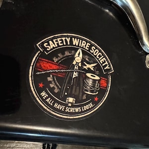 Può includere: Un adesivo circolare con la scritta "SAFETY WIRE SOCIETY" in un banner. L'adesivo presenta un design rosso e nero con strumenti, una bobina di filo e la frase "WE ALL HAVE SCREWS LOOSE". L'adesivo è su una superficie nera.