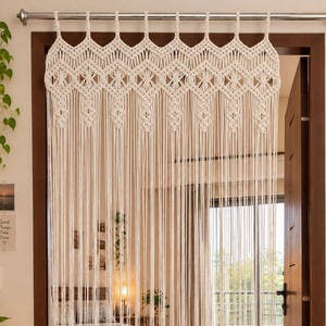 Op de afbeelding: Een gebroken wit macramé deurgordijn met een complex geknoopt chevron- en ruitpatroon aan de bovenzijde, dat overgaat in lange, rechte, verticaal hangende koorden die naar beneden vallen vanaf een zilveren metalen gordijnroede boven de deur.