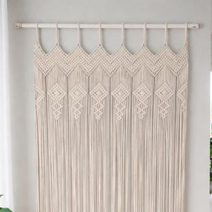 Puede incluir: Una cortina de macramé color hueso colgada de una barra de madera horizontal. La sección superior presenta un patrón de chevrón repetitivo, seguido de una serie de nudos en forma de diamante que terminan en largos flecos verticales.