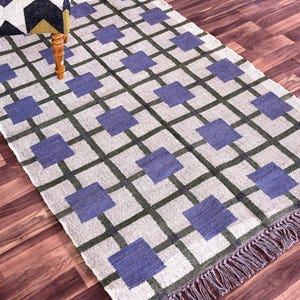 May include: A rectangular area rug with a geometric pattern. The rug features a grid of dark green lines with blue squares at the intersections. The background is a light beige color. The rug has a fringe on one end.