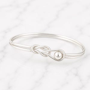 Könnte beinhalten: Ein silberfarbenes Armreif-Armband mit einem Knoten-Design und einem kleinen Kugelakzent. Das Armband besteht aus dünnem, rundem Metall und hat einen schlichten, eleganten Stil. Das Armband wird auf einer weißen Marmoroberfläche gezeigt.