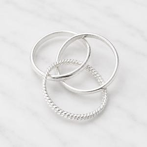 Könnte beinhalten: Drei silberne Ringe, die ineinandergreifen und ein einzigartiges Design bilden. Zwei Ringe haben eine glatte, polierte Oberfläche, während der dritte eine gedrehte Seiltextur aufweist. Die Ringe sind auf einer weißen Marmoroberfläche dargestellt.