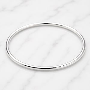 Könnte beinhalten: Ein einfacher, silberfarbener Armreif. Der runde Armreif ist glatt und glänzend und reflektiert das Licht. Der Schmuck ist vor einem weißen Marmorhintergrund platziert.