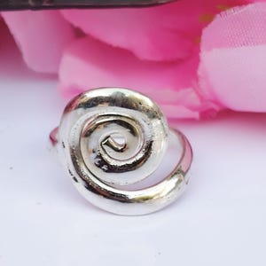 Könnte beinhalten: Ein silberner Ring mit einem Spiraldesign. Der Ring besteht aus poliertem Metall und weist ein wirbelndes Muster auf. Der Ring befindet sich auf einer weißen Oberfläche mit rosa Blumen im Hintergrund.
