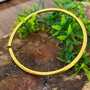 Könnte beinhalten: Ein glatter, kreisförmiger Armreif aus goldfarbenem Metall mit einer einzelnen Öffnung. Das Armband zeichnet sich durch eine polierte, reflektierende Oberfläche und ein schlichtes, durchgehend abgerundetes Design mit gleichbleibender Dicke aus.
