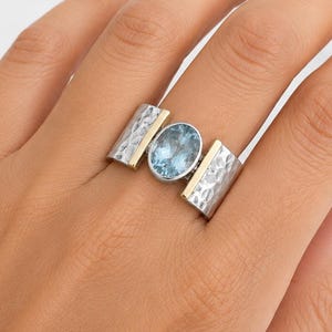 Könnte beinhalten: Ein silberner Ring mit einem hellblauen, ovalen Edelstein. Der Ring hat goldfarbene Akzente und ein strukturiertes Design an den Seiten. Der Ring befindet sich an einem Finger.