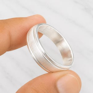 Könnte beinhalten: Ein silberner Ring mit gebürsteter Oberfläche und einem gerillten Mittelband. Der Ring wird zwischen zwei Fingern gehalten, was sein schlichtes Design hervorhebt. Die Oberfläche des Rings reflektiert das Licht und unterstreicht seinen metallischen Glanz.