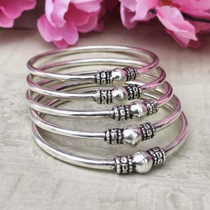 Könnte beinhalten: Eine Ansammlung silberner Armreifen mit dekorativen Perlenverzierungen. Jeder Armreif hat eine zentrale, runde Perle, die von detaillierten, gemusterten Metallarbeiten flankiert wird. Die Armreifen werden vor einem weichen, neutralen Hintergrund mit rosa Blüten im Hintergrund präsentiert.