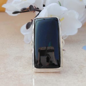 Könnte beinhalten: Rechteckiger Ring aus schwarzem Onyx mit polierter Oberfläche, gefasst in einem silberfarbenen Metall. Der Ring wird vor einem weichen, neutralen Hintergrund mit weißen Blumen präsentiert.