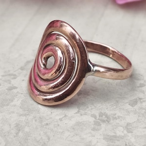 Könnte beinhalten: Ein kupferfarbener Ring mit einem Spiraldesign. Der Ring hat eine große, ovale Oberfläche mit einem erhabenen, wirbelnden Muster. Das Band ist ein einfaches, abgerundetes Design. Der Ring besteht wahrscheinlich aus Kupfer oder einer Kupferlegierung.