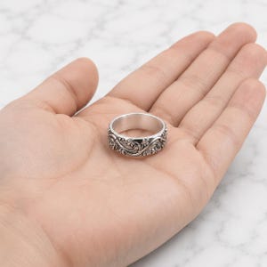 Könnte beinhalten: Ein silberfarbener Metallring mit einem detaillierten, geprägten Blumen- und Rankenmuster, das sich um das gesamte Band zieht. Das Design zeigt kleine, ausgearbeitete Blüten und geschwungene Blattmotive mit dunklen Vertiefungen zur Akzentuierung.