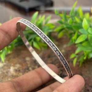 Könnte beinhalten: Ein dünner silberfarbener Armreif mit einem durchgehenden geprägten Muster aus kleinen, sich wiederholenden Blumenmotiven auf einem dunklen, vertieften Hintergrund. Die Metalloberfläche ist an den Kanten des Reifs poliert.
