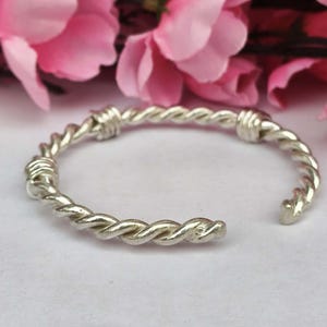 Könnte beinhalten: Ein silberfarbenes, offenes Armband mit einem gedrehten Seildesign. Das Armband besteht aus glänzendem Metall und befindet sich vor einem weichen, weißen Hintergrund mit rosa Blumen im Hintergrund.