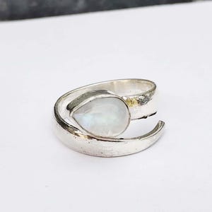 Könnte beinhalten: Ein silberner Ring mit einem birnenförmigen, irisierenden, weißen Edelstein. Der Ring hat eine strukturierte, gehämmert wirkende Oberfläche und umschließt den Finger in einem offenen Design. Der Edelstein ist der Blickfang und fängt das Licht ein.