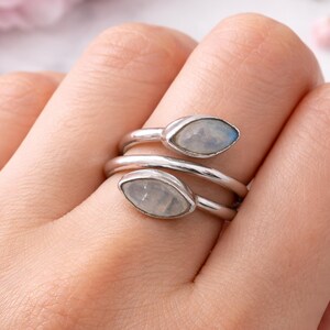Könnte beinhalten: Ein silberfarbener Ring mit Spiraldesign, besetzt mit zwei ovalen Mondstein-Edelsteinen. Die Edelsteine haben ein milchiges, schillerndes Aussehen. Der Ring wird an einem Finger getragen.