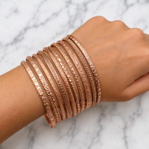 Könnte beinhalten: Ein Set aus zehn Armreifen aus roségoldfarbenem Metall, die zusammen getragen werden. Jeder Armreif weist ein eigenes geprägtes Muster auf, darunter Herzformen, florale Motive, geometrische Linien und gedrehte Seilstrukturen für einen Lagenlook.