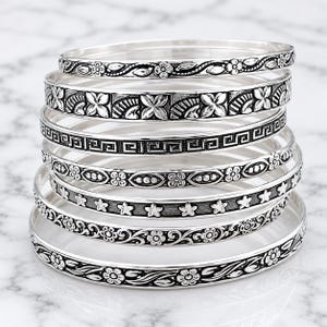 Könnte beinhalten: Ein Stapel aus sieben silberfarbenen Metallarmreifen, die jeweils mit unterschiedlichen geprägten Mustern wie Blumenmotiven, geometrischen griechischen Mäandern und Sternformen versehen sind. Die oxidierte Oberfläche betont die Details.