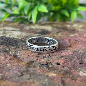 May include: A silver-toned metal ring featuring a band with engraved black Tibetan script characters circling the entire exterior surface. The ring has a smooth, polished finish and a consistent width, showcasing detailed dark lettering against the metal.