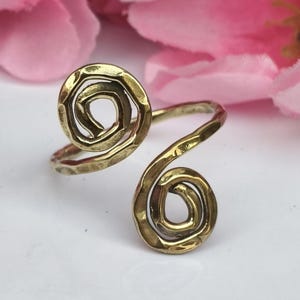 Könnte beinhalten: Ein messingfarbener Ring mit Spiralmuster. Der Ring hat zwei Spiralformen, die durch ein gebogenes Band verbunden sind. Das Metall hat eine gehämmerten Textur, und der Ring liegt auf einer weißen Oberfläche mit rosa Blumen im Hintergrund.