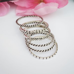 Könnte beinhalten: Eine Ansammlung von Silberringen mit verschiedenen Designs, darunter gedrehte Seile und geometrische Muster. Die Ringe sind vor einem weichen, weißen Hintergrund mit einer rosa Blume in der Ecke angeordnet. Diese Ringe eignen sich perfekt zum Kombinieren und verleihen einen Hauch von Eleganz.