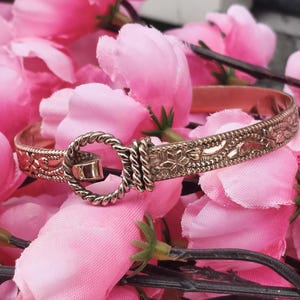 Könnte beinhalten: Ein goldfarbenes Armreif-Armband mit floralen und geometrischen Mustern. Das Armband hat einen seilartigen Verschluss und wird auf einem Bett aus rosa Blüten präsentiert. Das Design des Armbands umfasst detaillierte Gravuren.