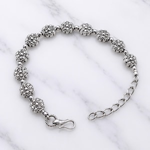 Könnte beinhalten: Ein silberfarbenes Armband mit einer Reihe sich wiederholender blumenförmiger Metallperlen, die durch kleine kugelförmige Abstandshalter verbunden sind. Das Armband verfügt über einen Metallhakenverschluss und eine verstellbare Kettengliedverlängerung.