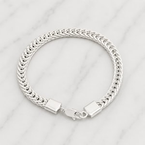 Könnte beinhalten: Ein silberfarbenes Armband mit einem geflochtenen Kettenmuster. Das Armband verfügt über einen sicheren Verschluss und wird auf einer weißen Marmoroberfläche präsentiert. Die Kettenglieder sind eng miteinander verbunden.
