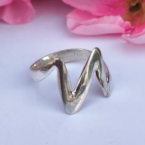May include: A silver ring with a unique zigzag design. The ring has a polished finish and a smooth, curved shape. The ring is set against a white background with pink flowers in the background. Jewelry.