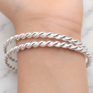Könnte beinhalten: Zwei polierte Silberarmreifen mit einem durchgehenden gedrehten Seildesign. Die Armbänder haben eine helle, reflektierende metallische Oberfläche und eine gleichmäßige kreisförmige Form, die zusammen getragen einen geschichteten Look erzeugen.