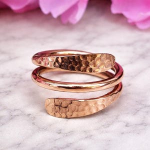 Könnte beinhalten: Ein roségoldfarbener Wickelring mit gehämmerten Enden. Der Ring hat ein spiralförmiges Design mit zwei vollständigen Schlaufen. Der Hintergrund ist eine Marmoroberfläche mit rosa Blüten.