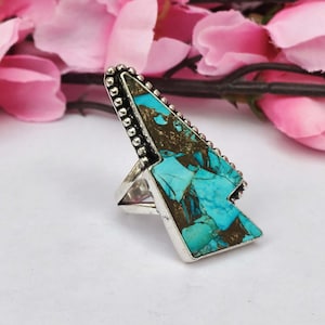 Op de afbeelding: Zilveren ring met een grote, turquoise steen. De steen heeft een uniek patroon van blauwe en bruine tinten. Het ontwerp van de ring is een pijlvorm, met een rand van kralen. De ring staat tegen een zachte roze achtergrond.