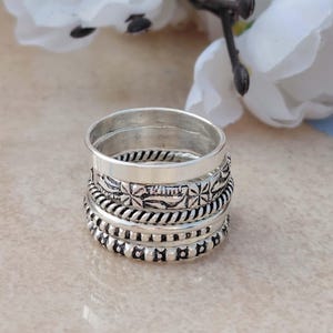 Könnte beinhalten: Ein Stapel silberner Ringe mit verschiedenen Texturen und Designs. Die Ringe umfassen glatte Bänder, gedrehte Seilmuster sowie detaillierte Blumen- und Perlenelemente. Die Ringe werden auf einer hellen Oberfläche mit weißen Blumen im Hintergrund präsentiert.