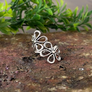 Könnte beinhalten: Ein silberfarbener Metallring mit zwei durchbrochenen Blumenmotiven im Bypass-Stil. Jede Blume besteht aus fünf tropfenförmigen Blütenblättern, die von einem zentralen runden Punkt ausgehen, mit polierter Oberfläche und dunklen Vertiefungen.