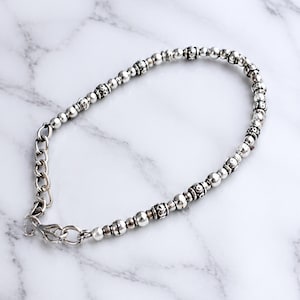 Könnte beinhalten: Ein silberfarbenes Perlenarmband mit abwechselnd glatten, runden Perlen und strukturierten, gemusterten zylindrischen Perlen. Das Armband verfügt über eine silberfarbene Metall-Verlängerungskette und einen Karabinerverschluss zum sicheren Schließen.