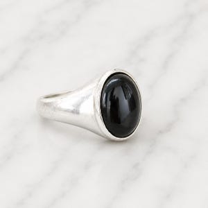 Könnte beinhalten: Ein silberner Ring mit einem großen, ovalen, schwarzen Stein. Der Ring hat ein schlichtes, klassisches Design mit poliertem Finish. Der schwarze Stein ist glatt und reflektierend, gefasst in einer Lünettenfassung.