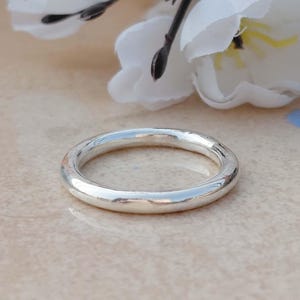Könnte beinhalten: Ein polierter Silberring mit abgerundetem Design. Der Ring reflektiert Licht und scheint aus Metall zu sein. Der Ring ist ein einfaches Band, das für den Alltag geeignet ist. Der Hintergrund umfasst weiße Blumen. Der Ring hat einen Durchmesser von ca. 2 cm.
