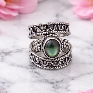 Könnte beinhalten: Zwei silberne Ringe mit aufwendigen Mustern. Der obere Ring hat ein wellenförmiges Design, während der untere Ring einen grünen ovalen Edelstein aufweist. Beide Ringe haben einen geflochtenen Rand. Die Ringe werden auf einer Marmoroberfläche präsentiert.