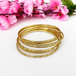 Könnte beinhalten: Ein Set aus vier gestapelten Armreifen aus goldfarbenem Metall. Jeder Armreif weist eine eigene Oberflächenstruktur auf, darunter florale Gravuren, Perlenverzierungen und glatt polierte Bänder, die ein geschichtetes goldenes Aussehen erzeugen.