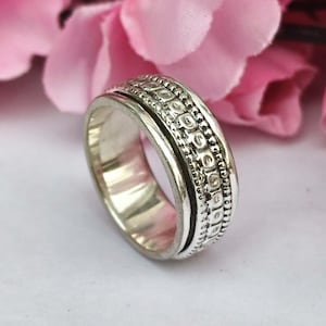Könnte beinhalten: Ein silberfarbener Ring mit einem strukturierten Band und einem drehbaren Außenring. Der Ring hat ein detailliertes Muster aus kleinen, erhabenen Punkten und Linien. Der Hintergrund zeigt zarte rosa Blüten.