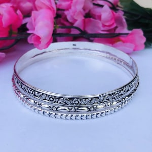 Könnte beinhalten: Ein silbernes Armreif-Armband mit aufwendigen Blumen- und Blättermustern. Das Armband hat ein detailliertes Design mit einer Reihe kleiner, sich wiederholender Formen an den Rändern. Rosa Blumen sind im Hintergrund.