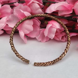 Könnte beinhalten: Ein offenes Armreif-Armband in Bronzefarbe mit einem gedrehten Seildesign. Das Armband ist vor einem Hintergrund aus rosa Blüten platziert. Der Durchmesser des Armbands beträgt etwa 6 cm.