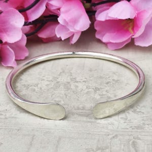 Könnte beinhalten: Ein silberfarbenes offenes Armreif-Armband. Das Armband hat ein glattes, abgerundetes Band, das sich zu flachen, tropfenförmigen Enden verjüngt. Rosa Blumen sind im Hintergrund.