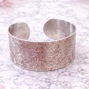 Könnte beinhalten: Ein breites, silbernes Armband mit einem komplizierten Blumenmuster. Das offene Armband hat eine strukturierte Oberfläche mit einem detaillierten Design aus wirbelnden Blättern und Blumen. Das Metall hat eine polierte, reflektierende Oberfläche, die ihm einen Vintage-Look verleiht.
