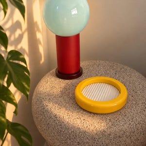May include: A table lamp featuring a spherical light blue glass shade atop a cylindrical red base, positioned next to a circular yellow tray with a white ribbed interior surface. The lamp and tray are arranged on a textured, speckled gray surface.