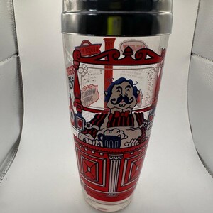 Pode incluir: Uma coqueteleira vintage com corpo transparente e tampa preta e prateada. A coqueteleira apresenta um design vermelho, branco e azul com uma ilustra&ccedil;&atilde;o de desenho animado de um homem de bigode e o texto "Beer 5&cent;".